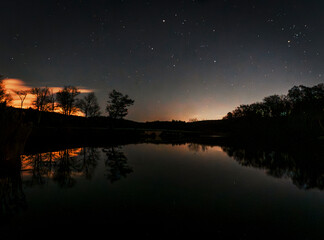 A star-filled night sky glows above Lake Kanawauke at Harriman State Park, where silhouetted trees and warm clouds reflect on the calm, glassy water, creating a serene, moody landscape.