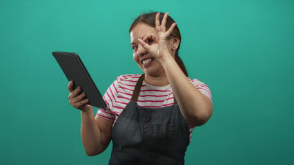 Woman holding tablet and making ok sign while wearing striped shirt and denim apron in teal studio; confidence learning tips.