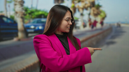 Woman in a pink jacket pointing happily on a sunny outdoor street with palm trees and blurred pedestrians in the background, embodying vibrant urban life and style.