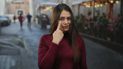 Fototapeta premium Woman pointing to her eye on a bustling terrace with blurred street background showcasing lively outdoor atmosphere in vibrant city setting.