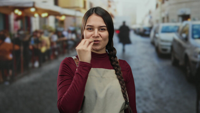 Young hispanic waitress in apron blows a kiss on a bustling restaurant terrace on a lively street surrounded by coffee drinkers and outdoor ambiance. - Powered by Adobe