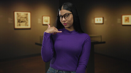 Woman with glasses and purple sweater makes call me gesture in gallery building; playful invitation.