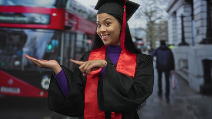 Woman in graduation gown pointing finger to palm on busy city street; academic achievement pride.