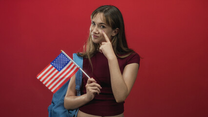 Young woman holding usa flag pointing finger to cheek in red studio; patriotism pride student youth.