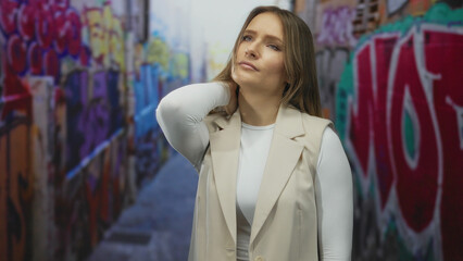 Naklejka premium Woman standing outdoors on city street with colorful graffiti holding neck in discomfort, wearing beige jacket, suggesting urban lifestyle.