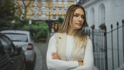 Fototapeta premium Woman standing confidently on a city street with crossed arms, blonde hair flowing and wearing a stylish beige vest, surrounded by parked cars and urban architecture.