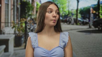 Fototapeta premium Teenager girl puckers lips and tilts head slightly in a light blue top with exposed shoulders on a sunny urban street scene; playfulness.