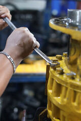 Close-up of a mechanic's hands using a combination wrench to manually tighten or loosen a series of large bolts on a heavy-duty yellow transmission or final drive casing during repair.