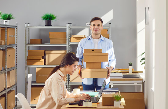 Young happy man employee in warehouse holding cardboard boxes. Woman working on laptop checking stock and inventory with male coworker. Small business owner preparing order for client in storeroom.