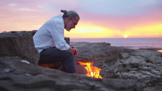 A contemplative man sits by a small fire on rocky terrain during a serene sunset by the sea. The scene evokes themes of mindfulness, reflection, and escaping daily life.