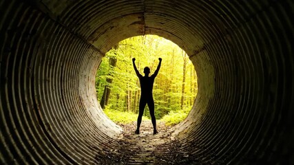 Silhouette of a person raising arms inside a dark metallic tunnel, with a bright green foliage background. Light and shadow play enhance the sense of escape and freedom.