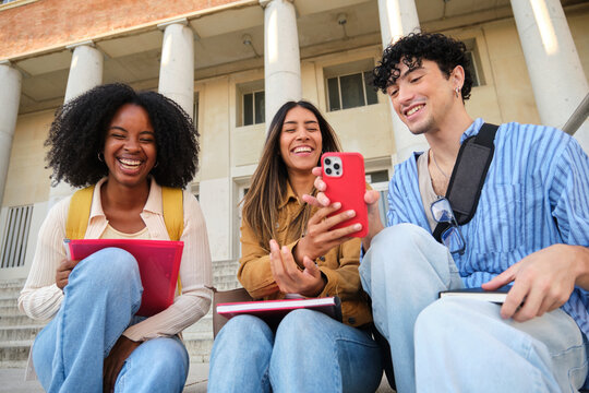 Group of happy students using a smartphone and laughing together on university steps - Powered by Adobe