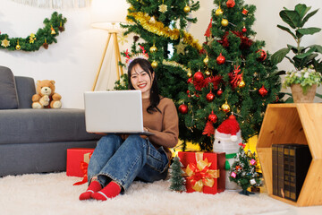 Asian young beautiful woman smile sitting on carpet floor wear reindeer antler using laptop computer, presents ornaments decorated in living room, Merry christmas happy new year festival celebrated.