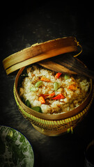 A rustic meal setup with a woven basket of seasoned rice, a vintage enamel plate with cutlery, and a cup of black coffee on a dark table. Warm lighting creates a homely, traditional Indonesian dining.