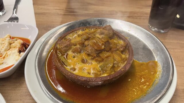 Lamb stew in a hot casserole dish, a steaming, spicy, delicious Turkish dish. Dipping bread by hand. High-angle shot.
