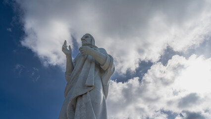 A huge statue of Christ against a background of blue sky and clouds. Sculpture made of white Carrara marble. Bottom-up view. Cuba. Havana. Jesus of Nazareth