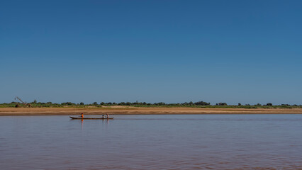 A traditional wooden pirogue boat with people sails along a red-brown river. Glare, reflection on the water. Green vegetation on a sandy coast. Clear blue sky. Copy space. Madagascar.Tsiribihina river