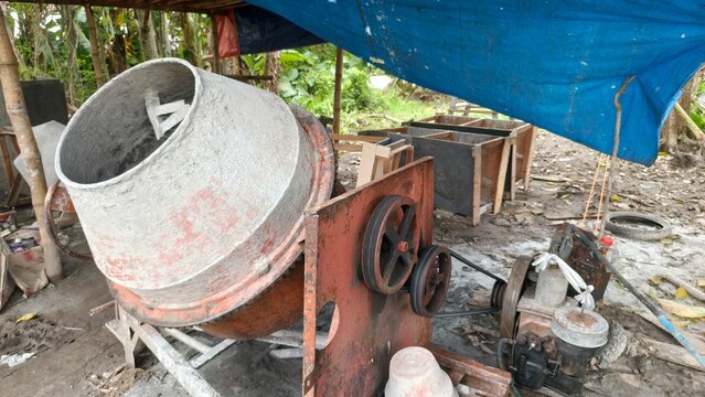 A worn concrete mixer machine in an outdoor construction area, surrounded by tools and materials under a blue tarp. This photo is perfect for content related to construction work, industry, building e