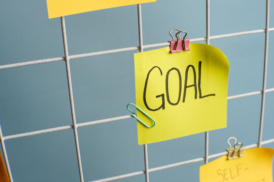 Yellow sticky note with the word “GOAL” attached to a metal grid board using a paper clip and binder clip. Motivational planning and productivity concept in a clean, minimal setup.