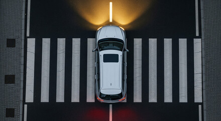 Aerial view of a white car on a crosswalk at night, with headlights illuminating the road and zebra stripes.