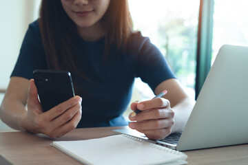 Young asian woman using smartphone and online working on laptop computer, surfing the internet at home office, digital nomad. Student studying online, remote work