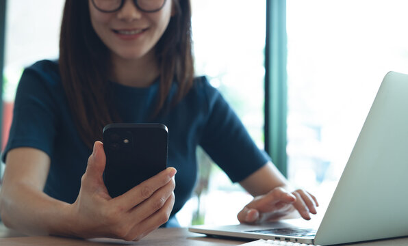 Woman remote working at cafe.  Happy young asian woman in casual clothes using smartphone and online working on laptop computer, surfing the internet at coffee shop or home office - Powered by Adobe