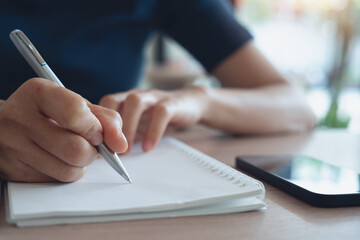 Closeup, woman hand with a pen writing on paper notebook with digital tablet on office table, business planning. Student doing homework, writing on notepad at home
