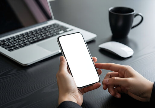Person holding a smartphone with a blank screen in front of a laptop and coffee mug