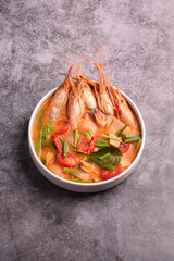 A bowl of authentic and spicy Thai Tom Yum Goong soup, featuring large river prawns, chili, and herbs, served against a dark, rustic background.