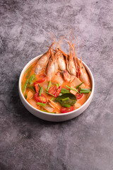 A bowl of authentic and spicy Thai Tom Yum Goong soup, featuring large river prawns, chili, and herbs, served against a dark, rustic background.