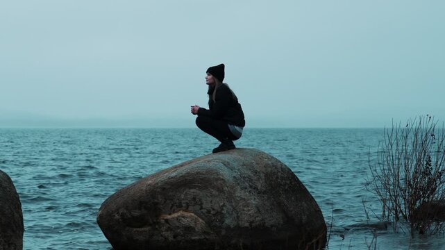 A lonely girl is sitting on a big stone on the shore of a lake on a cloudy rainy day and enjoying nature. Cool weather. The harmony of man and nature.