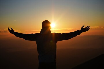 Fototapeta premium Silhouetted person with arms outstretched, standing on a hill at sunset/sunrise. Powerful image of freedom, spiritual connection, achievement, and natural beauty.