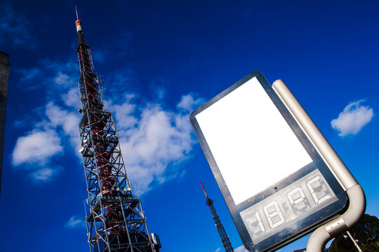 Blank advertising billboard and a tall telecommunication tower against a deep blue sky. Modern street sign with a digital clock, perfect for mockups about media, technology, and urban communication.