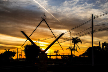 Silhouette of the Masonic Square and Compasses monument against a dramatic golden sunrise sky in a Brazilian city, symbolizing mystery, fraternity, and order