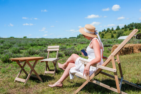 Young beautiful sexy woman in straw hat and sunglasses sitting on a sun lounger and works on a smartphone on a meadow on a sunny day against the sky. Rest. Vacation. Tourism. - Powered by Adobe