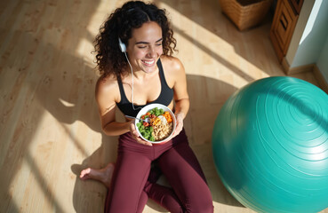 Young woman listens to music wearing earphones while eating healthy food bowl on floor. Fitness ball nearby suggests post workout meal or rest day.