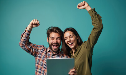 Happy couple cheers success with tablet. Man and woman celebrate victory raising their arms. Pair enjoy online win using digital device on blue background. Good news makes people happy.