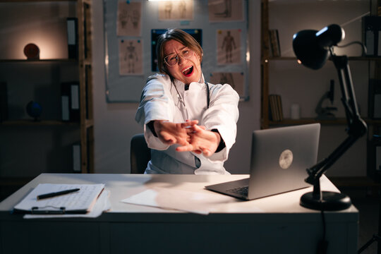 Lazy sleepy female doctor in eyeglasses stretching arms and yawning on workplace, tired and overworked