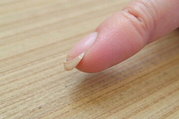 Natural fingernail with subtle imperfections, reflecting soft light on a textured, earthy surface. Macro view of a natural, unpolished fingernail. The surface shows subtle natural texture and slight