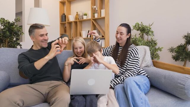 Parents and kids sitting together on sofa, making hand heart signs during a video call on laptop in bright cozy living room