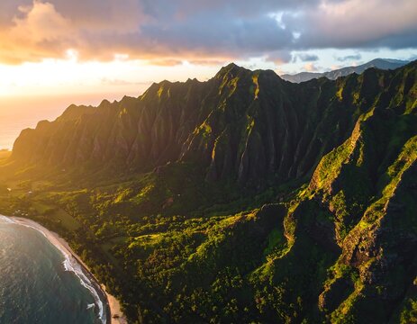 A vibrant sunset casts golden light on rugged mountains bordering a serene beach. The sky is filled with colorful clouds