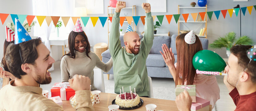 Joyful man raising hands while celebrating birthday with friends. Group of happy diverse people at desk wearing party hats cheering, clapping, enjoying cake and gifts in decorated living room. Banner. - Powered by Adobe