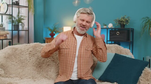 Caucasian mature man sitting on sofa at home, wearing wireless headphones and smiling. Middle-aged guy leans back, feeling rhythm of the music and gently dancing his hands, enjoying moment. Relaxing