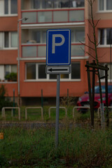 parking sign outside apartment building on grassy forecourt, bold blue P mounted on post with small placard, balcony windows and facade in background, trimmed lawn and hedge, quiet residential mood © Viktar