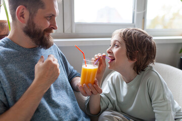 Bearded father and son drinking orange juice from one glass with two tubes. Man and boy laughing enjoying time together. Healthy morning routine, homemade juice, vitamin C, lifestyle.