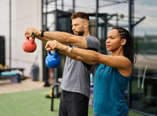 Portrait of a young black woman and white man coach or trainer exercising in a gym, lifting weights, kettlebell equipment, healthy lifestyle and strength exercise at fitness club concepts