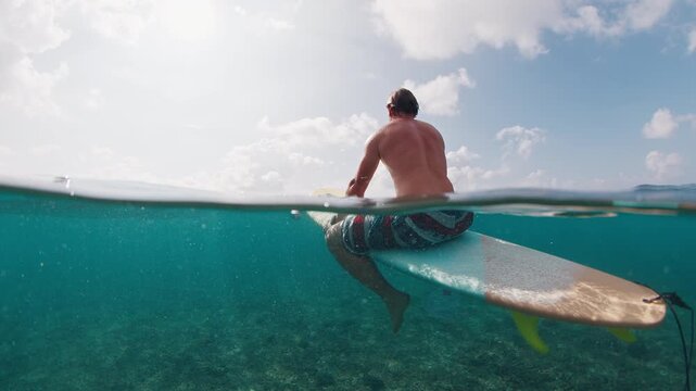 Surfer waits the wave. Underwater splitted view of the male surfer sitting in the ocean and waiting the wave