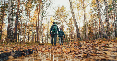 A father and son walking on the forest trail covered with fallen autumn leaves. The father-son relationship involves shared adventures that help strengthen their bond.