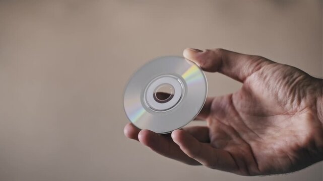 A male hand holds a small compact disc as light reflects off its iridescent surface, creating rainbow colors. This represents obsolete technology, data storage, and digital media from the 90s.