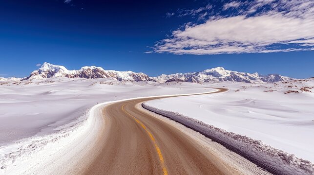 A winding road traverses a vast, snow-covered terrain with majestic, snow-capped mountains in the distance under a bright blue sky.